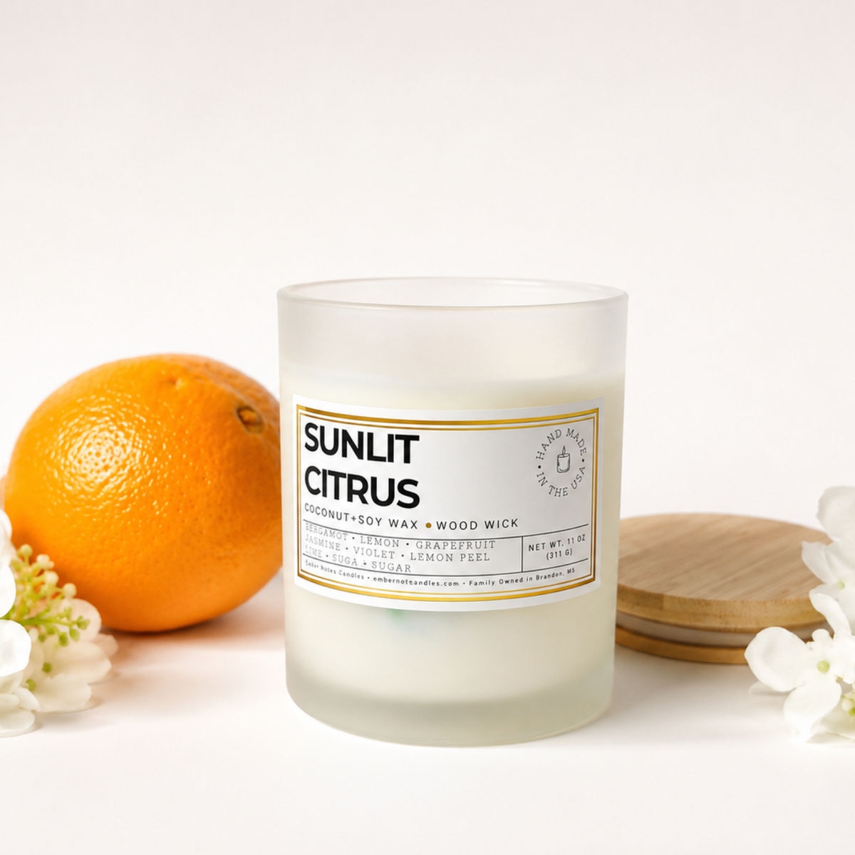 Sunlit Citrus coconut soy wax candle by Ember Notes Candles in a frosted white glass jar with a natural wood wick and gold-accented label, styled with a fresh orange, delicate white flowers, and a natural wooden lid against a bright white background.