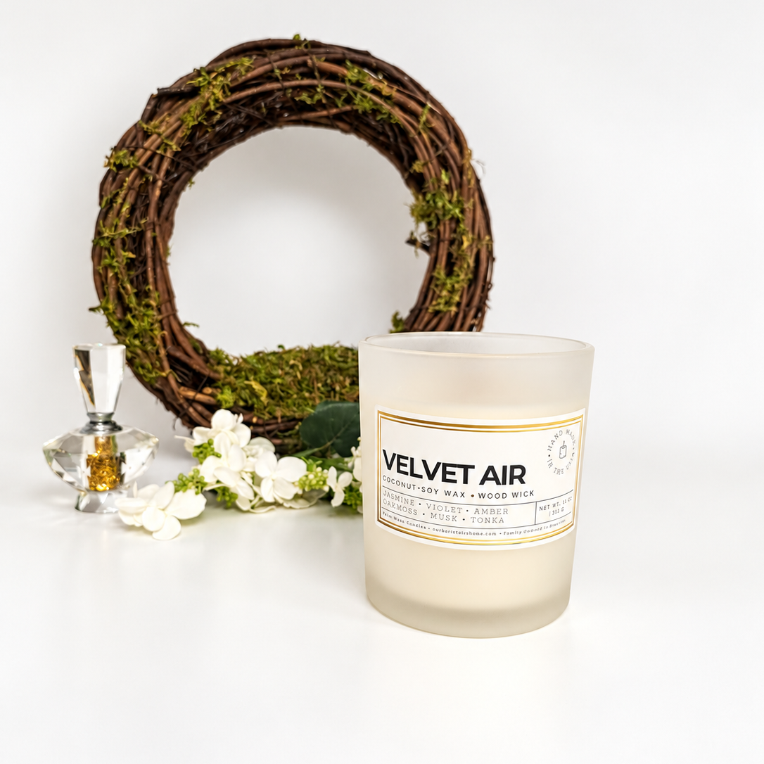 Angled view of the Velvet Air candle by Ember Notes Candles, featuring creamy coconut soy wax and a natural wooden wick in a frosted glass jar. Styled with white flowers, a moss-covered wreath, and a crystal bottle on a clean white background.