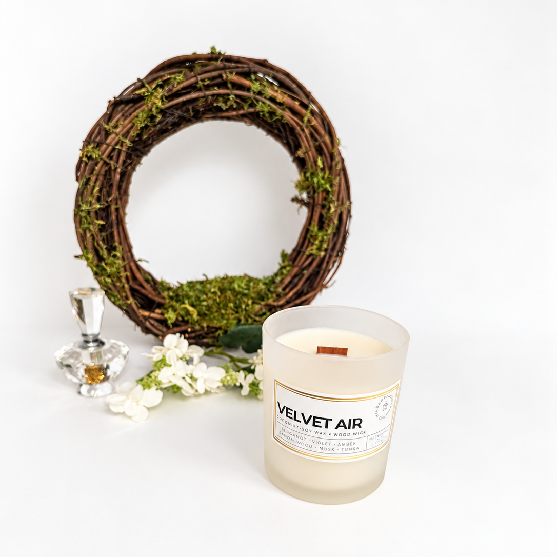 Velvet Air coconut soy wax candle by Ember Notes Candles in a frosted white glass jar with a gold-accented label, displayed with a moss-covered grapevine wreath, white flowers, and a decorative crystal bottle against a bright white background.